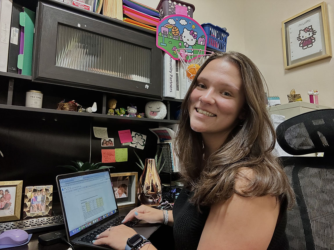 A college student with long dark hair sits at a laptop and smiles at the camera